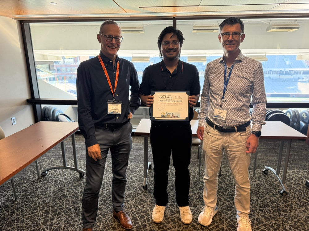Three men standing in a conference room overlooking a football stadium; the man in the center holds the Best Student Paper Award certificate.