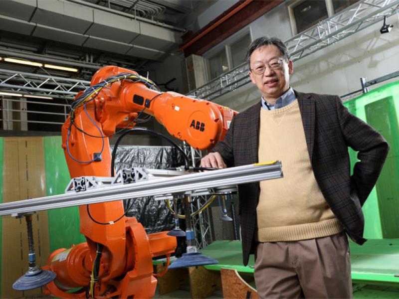 Asian man with black hair and glasses wearing a yellow sweater and black blazer standing next to an orange robot arm in a lab