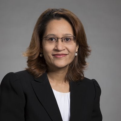 woman with brown shoulder length hair wearing glasses and a blazer on a grey background