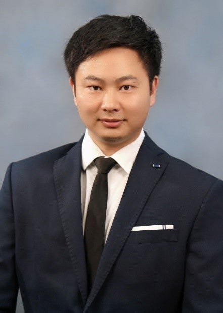 Headshot of East Asian man with black hair wearing a black suit and tie and white shirt on a blue background