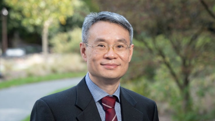 Headshot of an East Asian man with grey hair and glasses wearing a black suit, blue button up and red tie