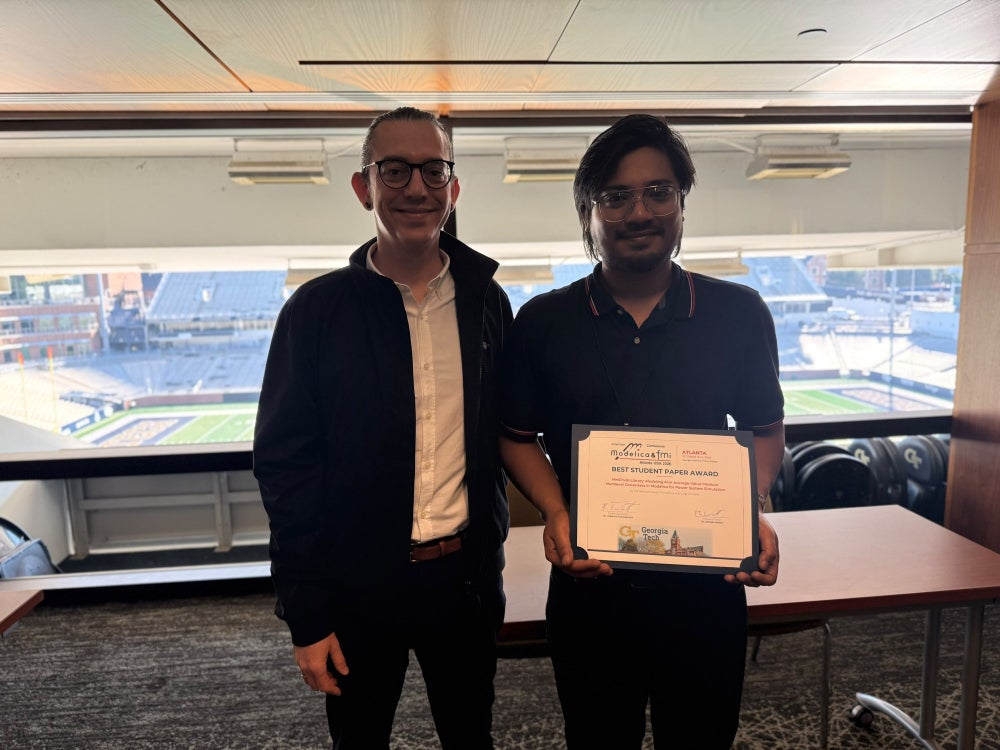 Two men standing in front of a window overlooking a football stadium; the man on the right holds an award certificate.